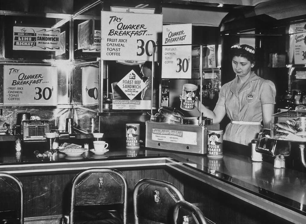 A woman in a diner uniform stands behind a counter, surrounded by vintage advertising posters for Quaker breakfast items and sandwiches. The decor features chrome and wood details typical of mid-20th-century diners.