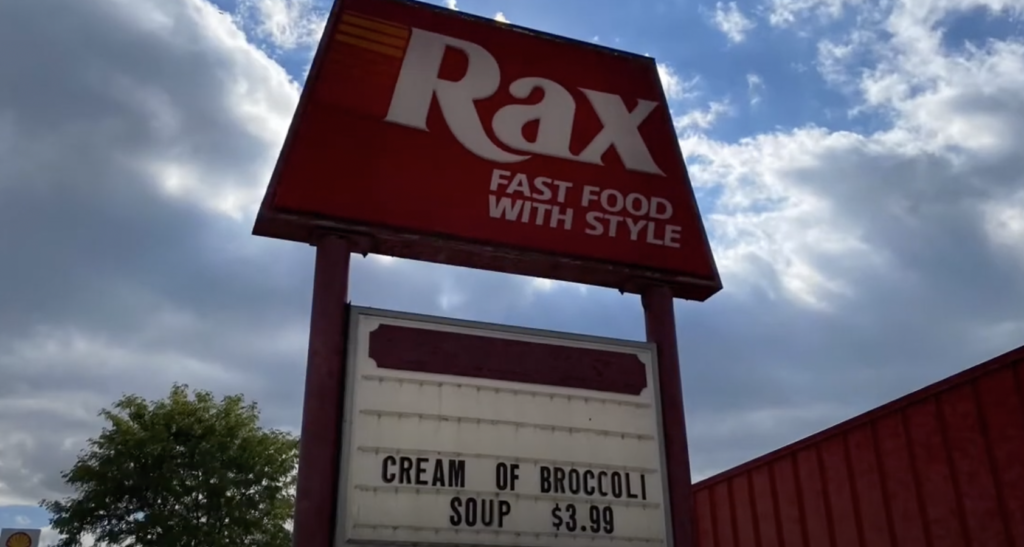 A red Rax fast food sign stands against a partly cloudy sky. Below, a white marquee reads "CREAM OF BROCCOLI SOUP $3.99." Trees are visible in the background.