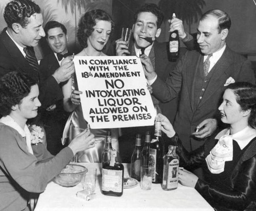 A group of people in a 1920s-style setting hold and look at a sign stating "In compliance with the 18th amendment, no intoxicating liquor allowed on the premises," while smiling and holding bottles of alcohol around a table.