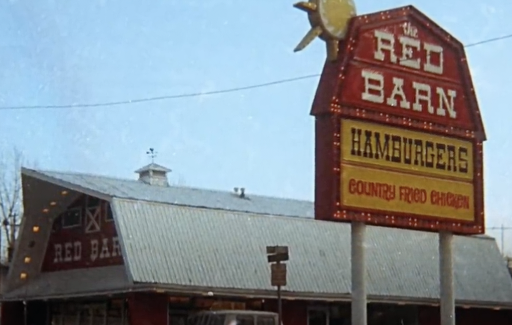 Vintage image of a Red Barn fast-food restaurant with a large red sign displaying "The Red Barn," "Hamburgers," and "Country Fried Chicken." The building has a barn-like appearance with a metal roof and barn-style windows.