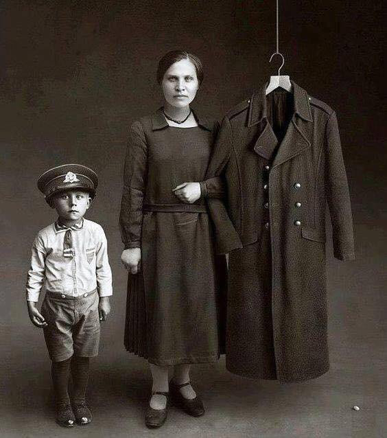 A solemn black and white photo shows a woman and a young boy standing beside a military coat hanging on a hanger. The boy wears a uniform, including a cap, while the woman holds the coat. The background is plain, emphasizing the trio.