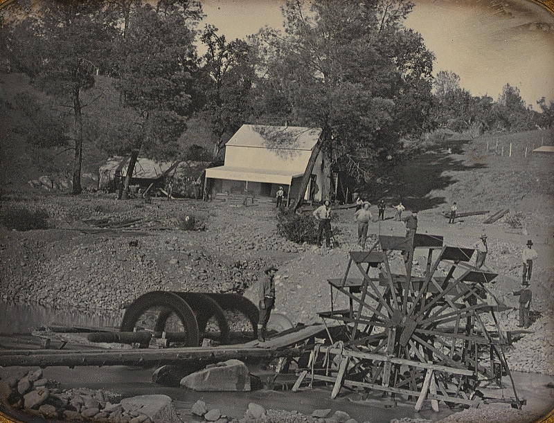 Sepia-toned vintage photograph of a rural scene with several people standing near a large wooden waterwheel by a river. A small building with a slanted roof is in the background, surrounded by trees. The landscape is rocky and sloped.