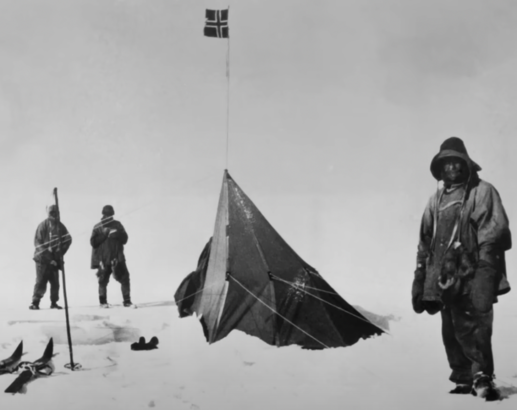 Three explorers stand near a tent in a snowy landscape. A flag is raised above the tent. The scene is black and white, suggesting a historical expedition in a cold environment.