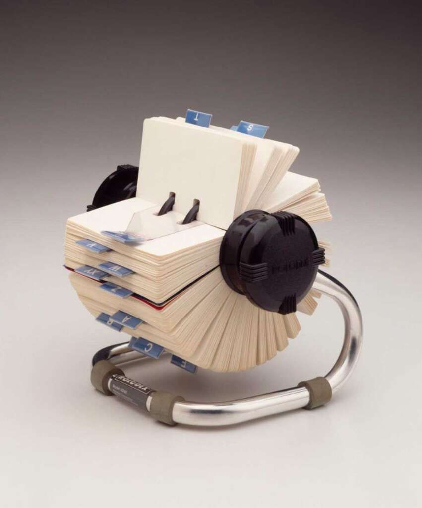 A vintage Rolodex file with a metal base and plastic knobs, holding a stack of white index cards organized by blue alphabetical tabs. The cards are slightly yellowed, indicating age.