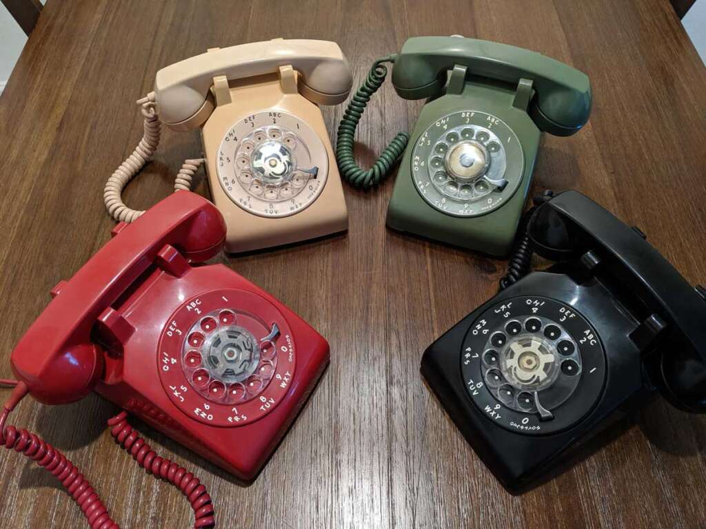 Four vintage rotary telephones are displayed on a wooden table. The phones are in different colors: cream, green, red, and black. Each phone has a coiled cord matching its color.