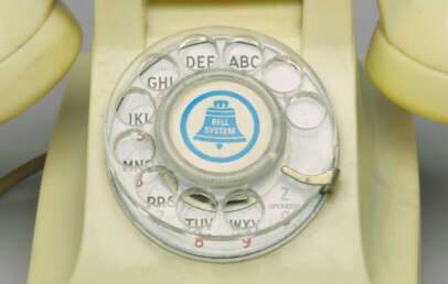 Close-up of an old-fashioned rotary dial telephone. The dial features letters and numbers with a central logo that says "Bell System." The phone is a yellowish color, and the dial has a metallic finger stopper.