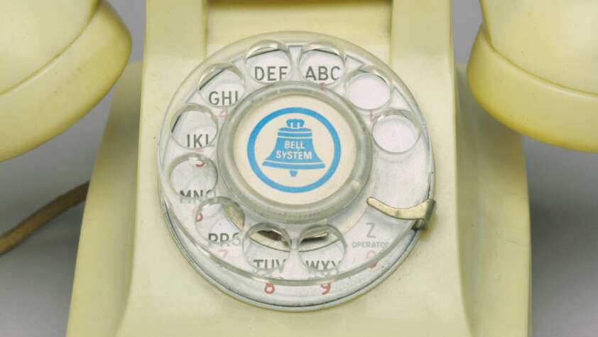 Close-up of an old-fashioned rotary dial telephone. The dial features letters and numbers with a central logo that says "Bell System." The phone is a yellowish color, and the dial has a metallic finger stopper.