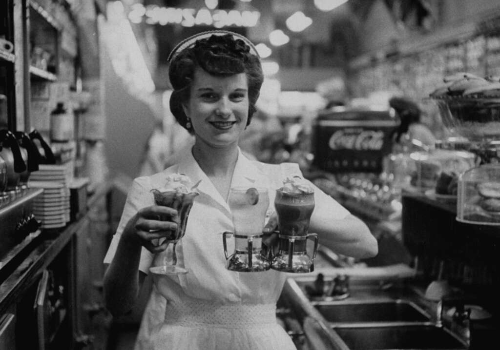 Black and white image of a smiling woman in a diner uniform holding a tray of ice cream sundaes and milkshakes. She stands behind a counter with Coca-Cola signage, dishes, and various diner items visible in the background.