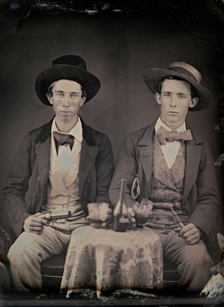 Two men in 19th-century attire sit side by side. Both wear dark jackets, bow ties, and brimmed hats. A small round table in front of them holds a bottle and two glasses. The image has a vintage, sepia-toned appearance.