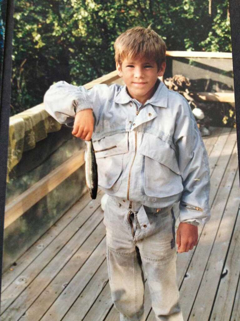 A young boy stands on a wooden deck, holding a fish by the mouth with his right hand. He is wearing a light-colored jacket and jeans. Behind him, there is a railing and green foliage.