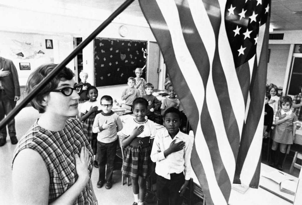 A classroom of diverse children and a teacher stand with hands over hearts, facing a large American flag. The room has a chalkboard and posters, and the children appear attentive. The setting is likely a pledge of allegiance or patriotic event.