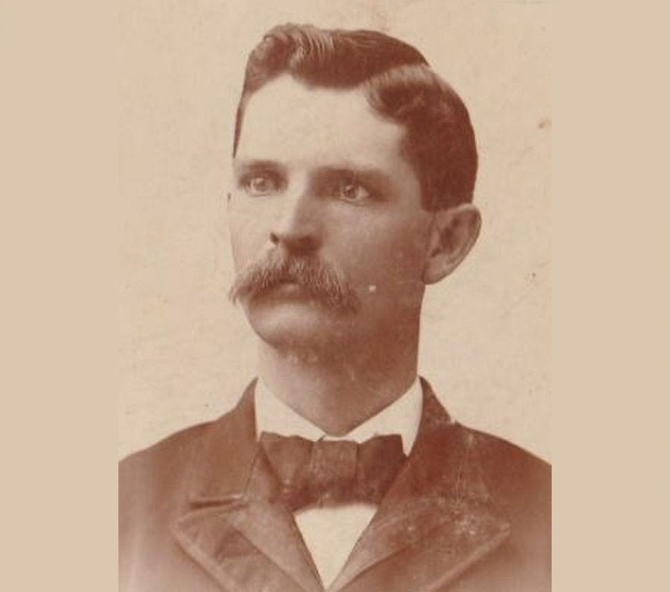 A sepia-toned portrait of a man with a well-groomed mustache and short hair. He is wearing a dark suit with a bow tie and a high-collared shirt. The background is plain, highlighting his formal attire and serious expression.