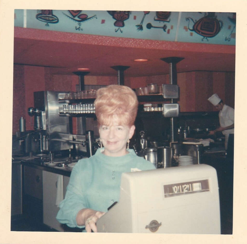 A woman with a tall, styled hairdo and a blue outfit stands smiling behind a cash register in a retro diner kitchen. The decor features vibrant patterns. In the background, a chef is visible working in the kitchen.