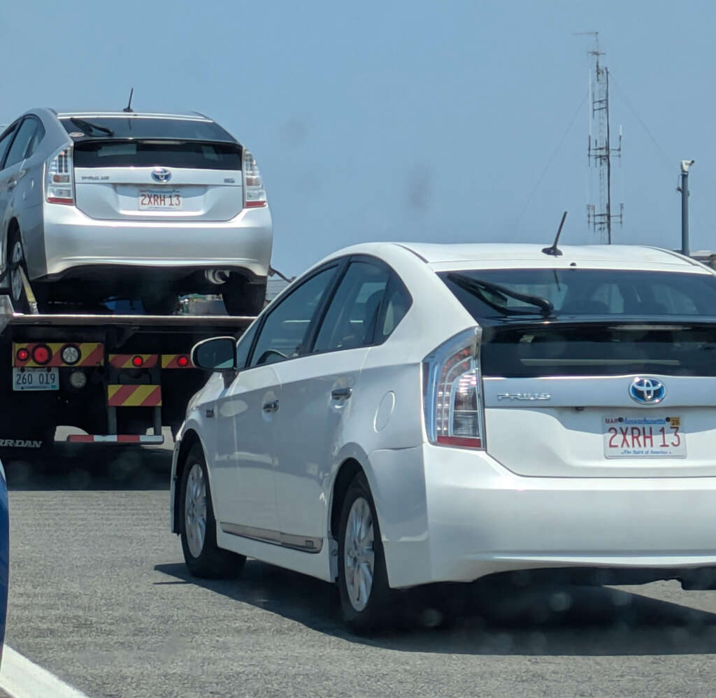 A silver car with a license plate "2XRH 13" is on a flatbed tow truck, while an identical white car with the same plate is driving behind it on the road. Clear blue sky in the background.