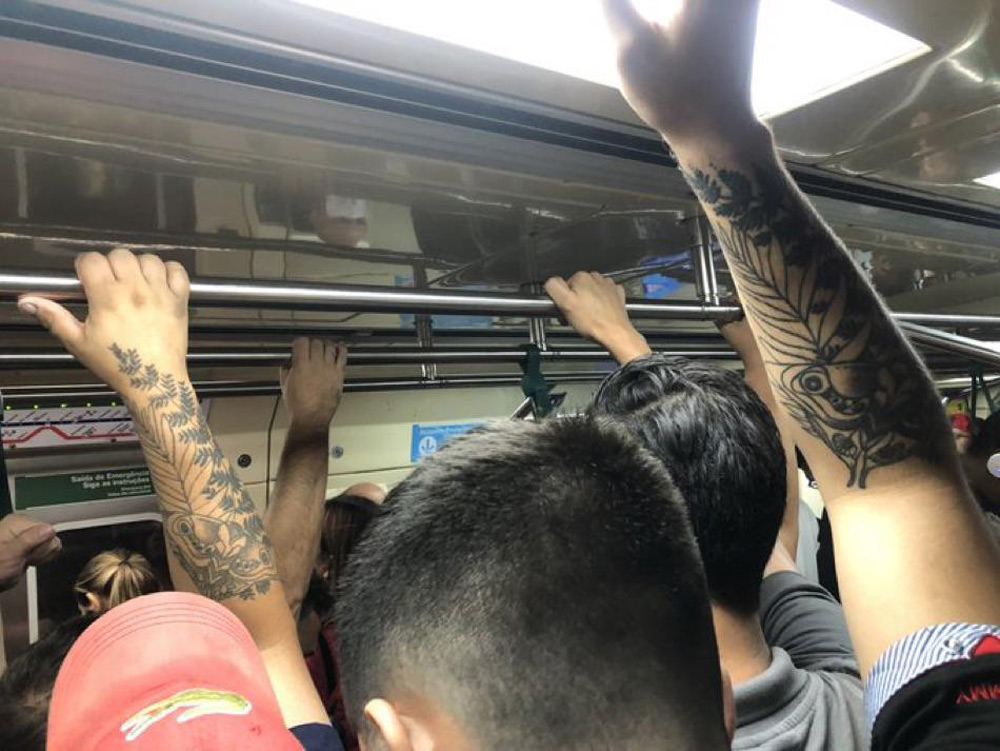 A crowded subway scene with people standing and holding onto overhead rails. Several arms have visible tattoos, including floral and owl designs. The setting is busy, with close proximity between passengers.