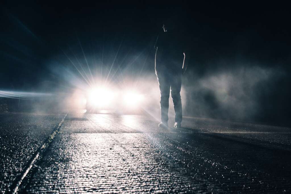 A person stands on a dark road facing the bright headlights of an approaching car, surrounded by mist. The scene is illuminated by the glare of the lights, creating a dramatic atmosphere.