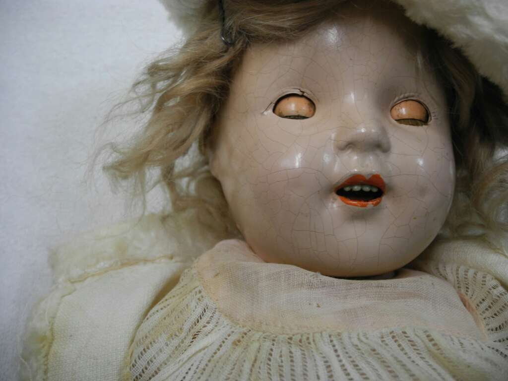 Close-up of an antique porcelain doll with cracked face paint, blonde curly hair, and partially closed eyes. The doll wears a cream-colored dress with lace detailing against a plain background.