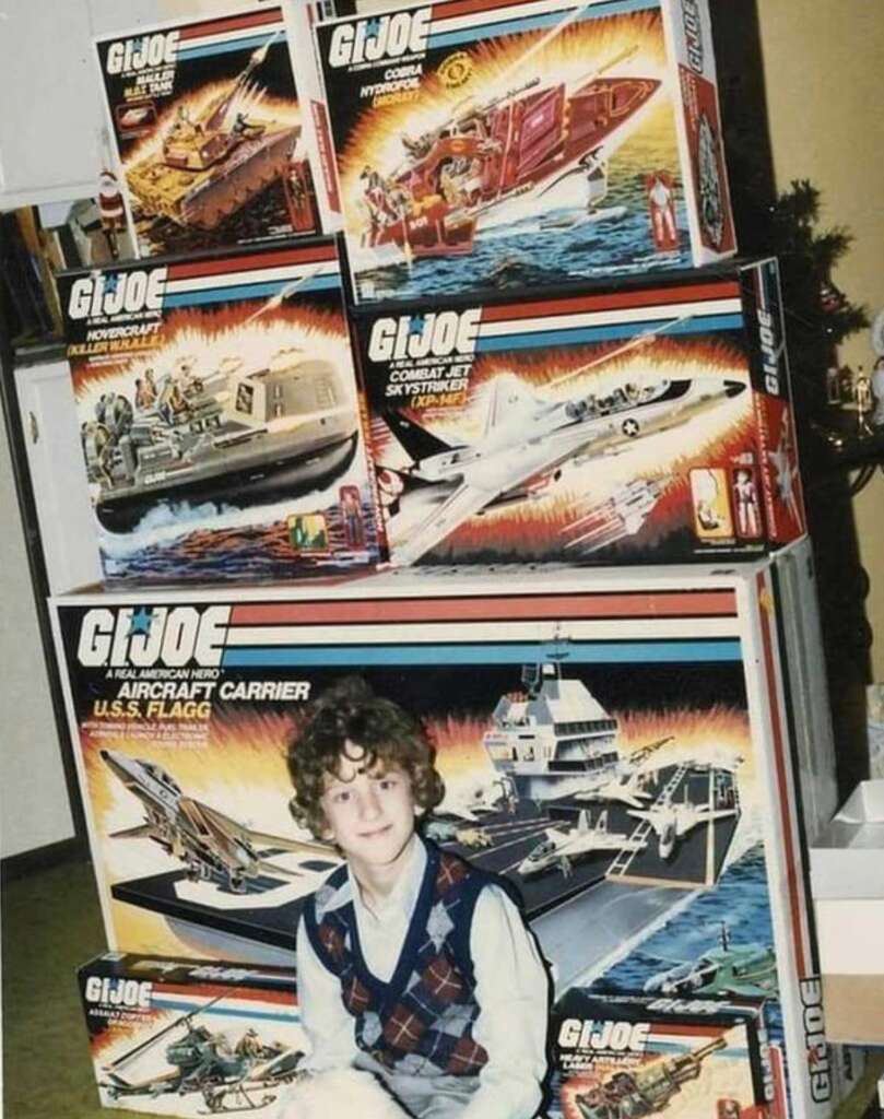 A child with curly hair sits in front of a tower of GI Joe toy boxes, including an aircraft carrier, combat jet, hovercraft, assault copter, and tank. The toys are arranged in a stack and the child is smiling.