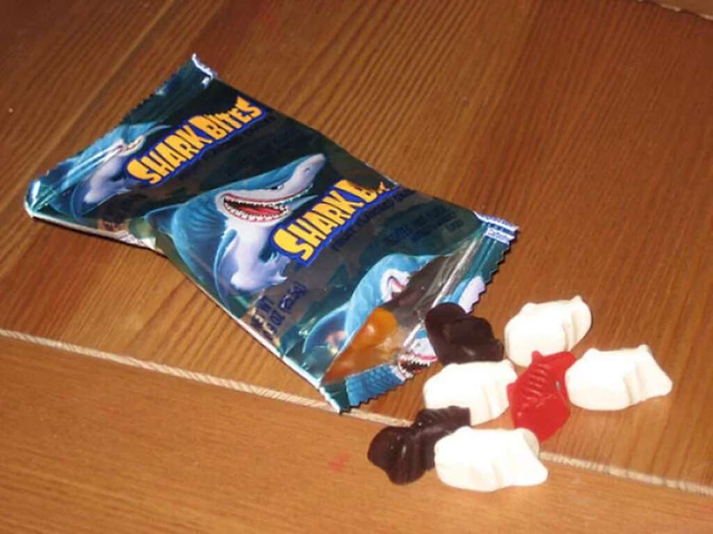 An open bag of "Shark Bites" gummy candies lies on a wooden surface. Several gummies in shark shapes are scattered around, including red, black, and white colors. The packaging features a cartoon shark graphic.