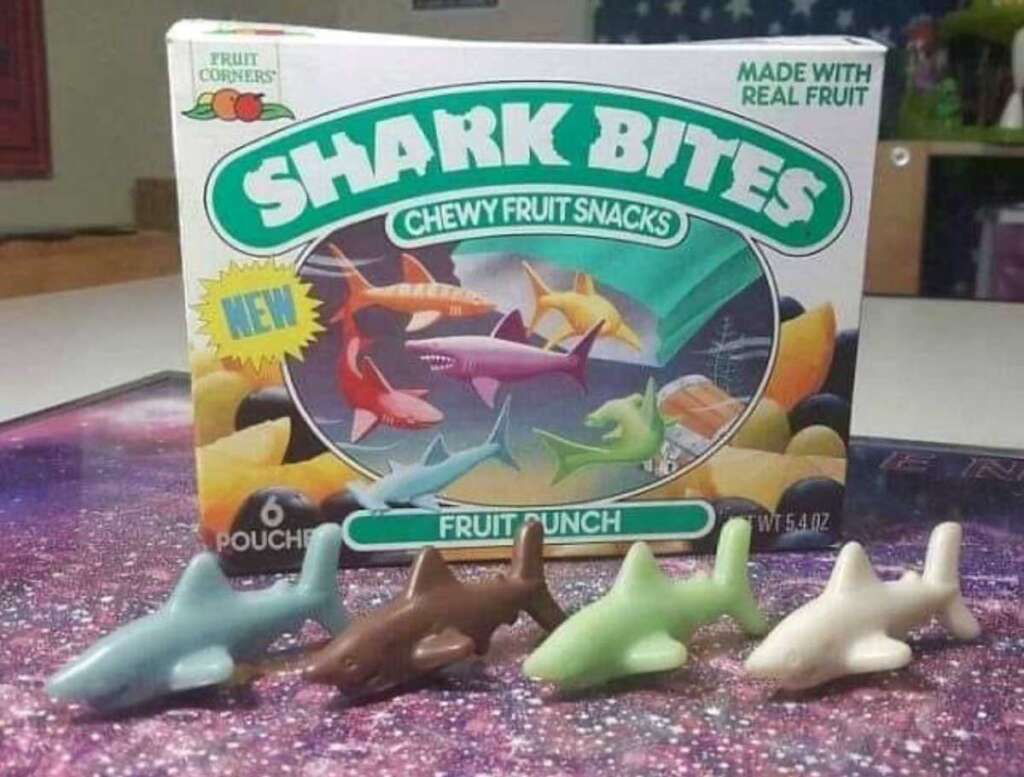 A box of Shark Bites chewy fruit snacks, featuring images of colorful sharks and labeled "Made with Real Fruit." In front are four shark-shaped gummies in blue, brown, green, and white. The flavor is fruit punch.