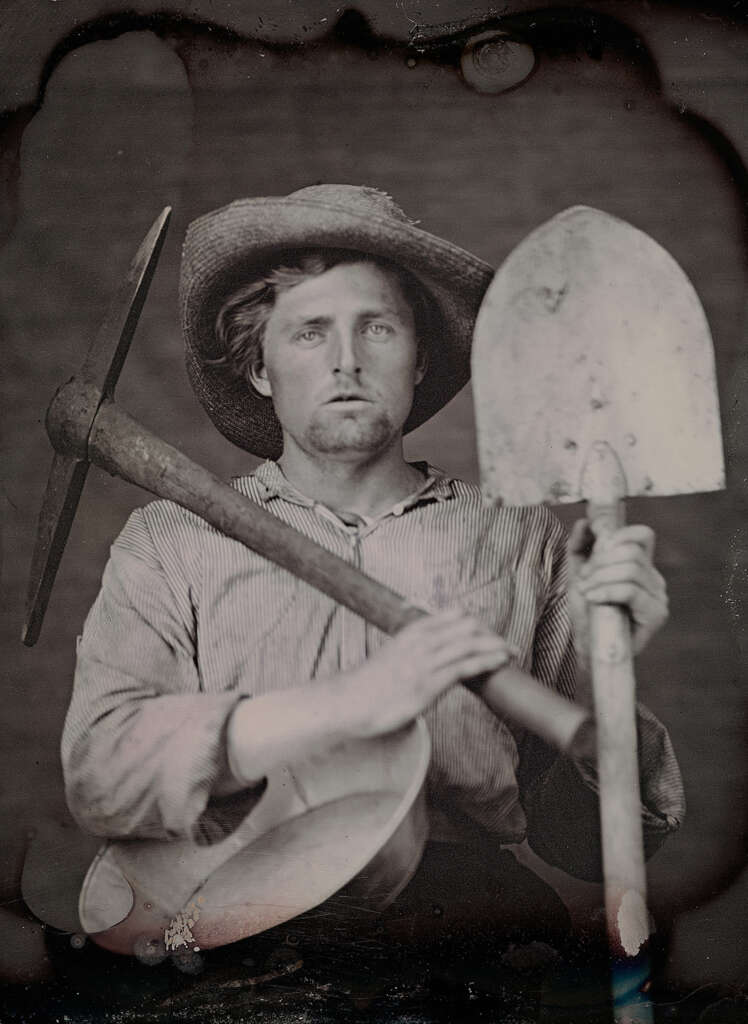 A vintage black and white photo shows a man wearing a striped shirt and straw hat, holding a shovel and pickaxe crossed in front of him. He has a serious expression and holds a pan in one hand, suggesting a mining theme.