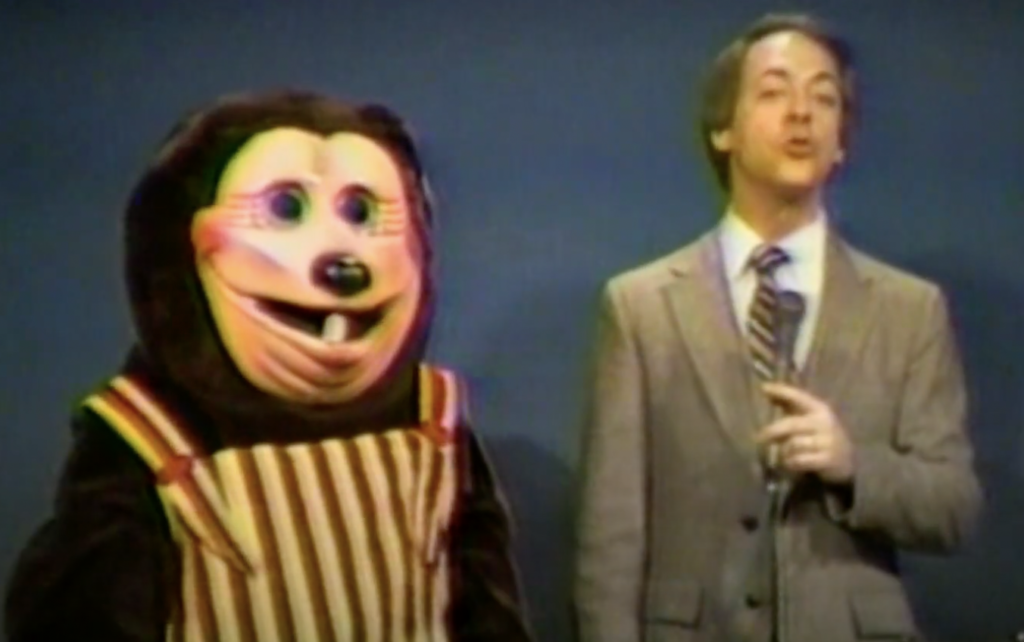 A person in a suit is speaking into a microphone on stage next to a mascot in a colorful costume with striped overalls and round facial features, set against a plain background.