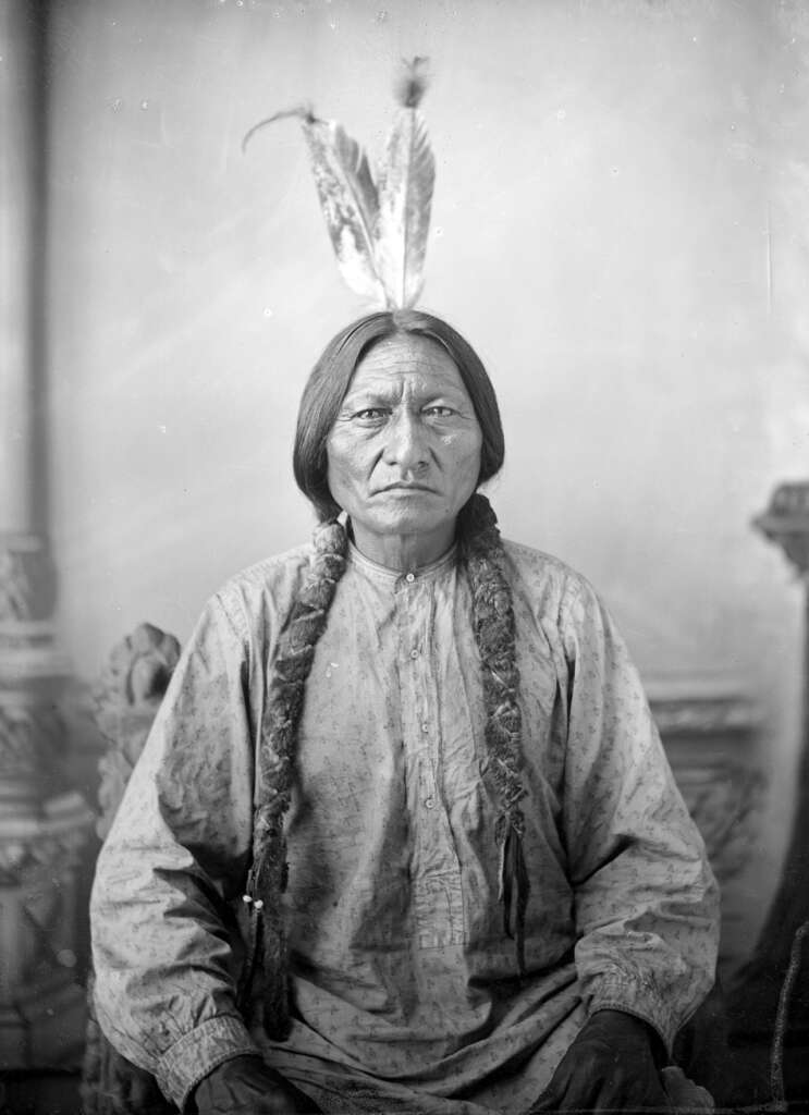 A person with long braids and feathers in their hair, wearing a traditional garment, is seated and facing forward. The background is plain, highlighting the subject's solemn expression.