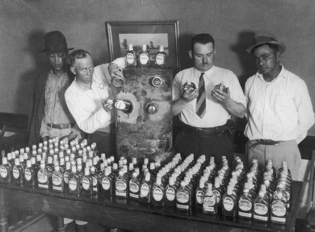 Four men stand behind a table filled with bottles labeled “Corno-malt syrup.” They examine the bottles and a metal contraption resembling a still. A framed picture hangs on the wall behind them. Black and white image.