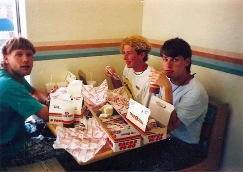 Three young men sitting at a Taco Bell table, surrounded by open taco wrappers and drink cups. They appear to be enjoying their meal and are engaged in conversation. The restaurant has a typical fast food interior with simple decor.