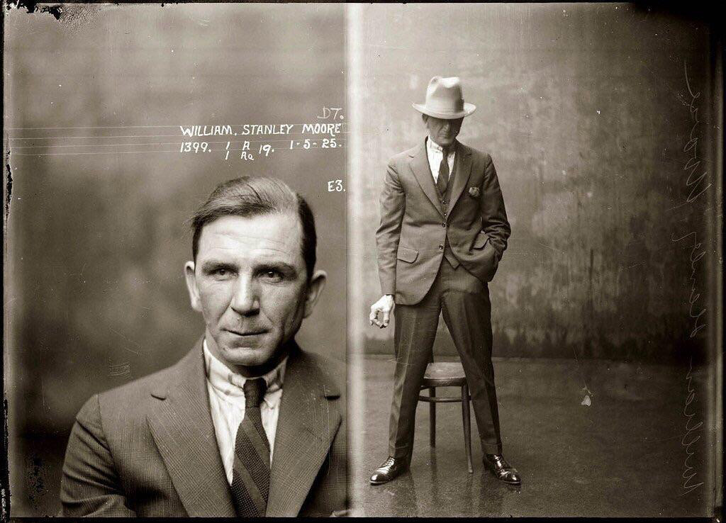 A vintage mugshot features two images of a man. On the left, he faces the camera in a suit and tie. On the right, he stands in a relaxed pose, leaning against a chair while holding a cigarette, wearing a suit and a fedora. Text is visible on the left.