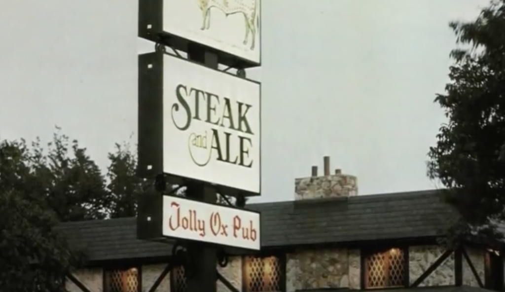 A vintage-style pub building with a sign displaying "Steak and Ale" and "Jolly Ox Pub." The structure has a stone exterior and wooden elements, surrounded by trees.