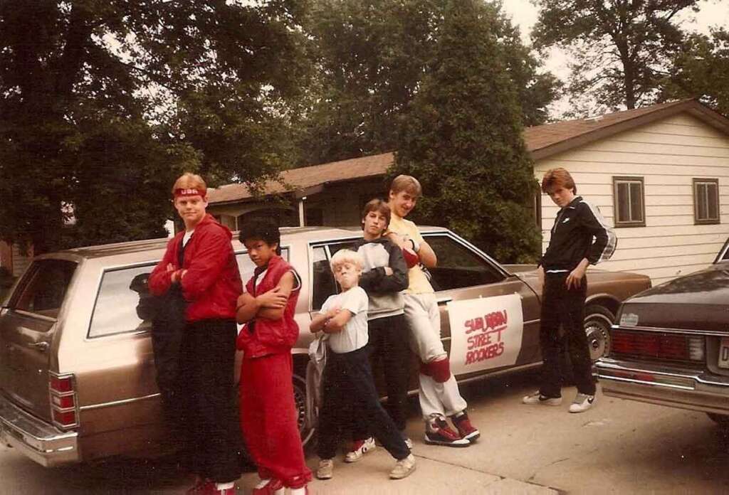 A group of six kids dressed in martial arts outfits stand confidently in front of an old station wagon with "Samurai Street Rockers" written on the side. Trees and a house are in the background.