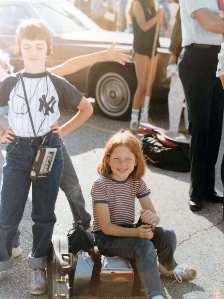 Two children in casual clothes are outdoors; one is standing with a Walkman and a Yankees shirt, the other is sitting and smiling on a toy car. People and a car are visible in the background. It's sunny, with long shadows on the ground.