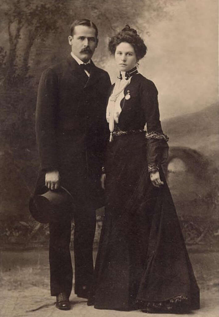 A vintage sepia-toned photograph of a man and a woman standing side by side. The man is wearing a dark suit and holding a hat, while the woman is dressed in a long dark gown with intricate details. A painted backdrop is behind them.