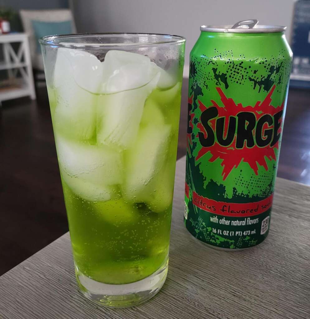 A can of Surge soda is placed next to a glass filled with ice and bright green Surge soda on a table. The can features a vibrant design with red and green colors. The background shows a blurred living room setting.