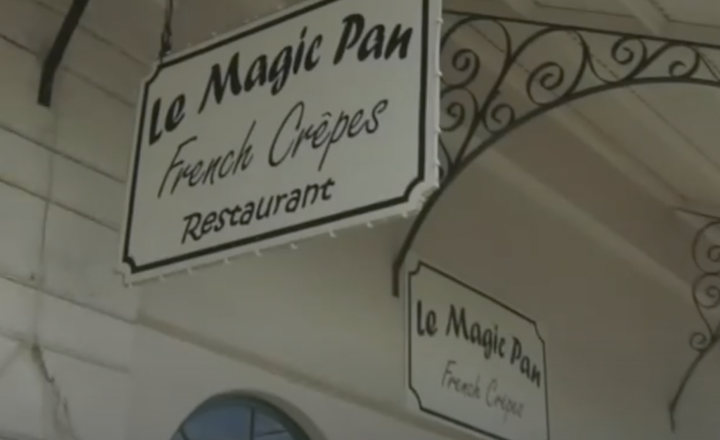 A vintage-style sign for "Le Magic Pan" hangs under an ornate wrought iron structure. The text reads, "Le Magic Pan French Crêpes Restaurant." Another similar sign is partially visible in the background.