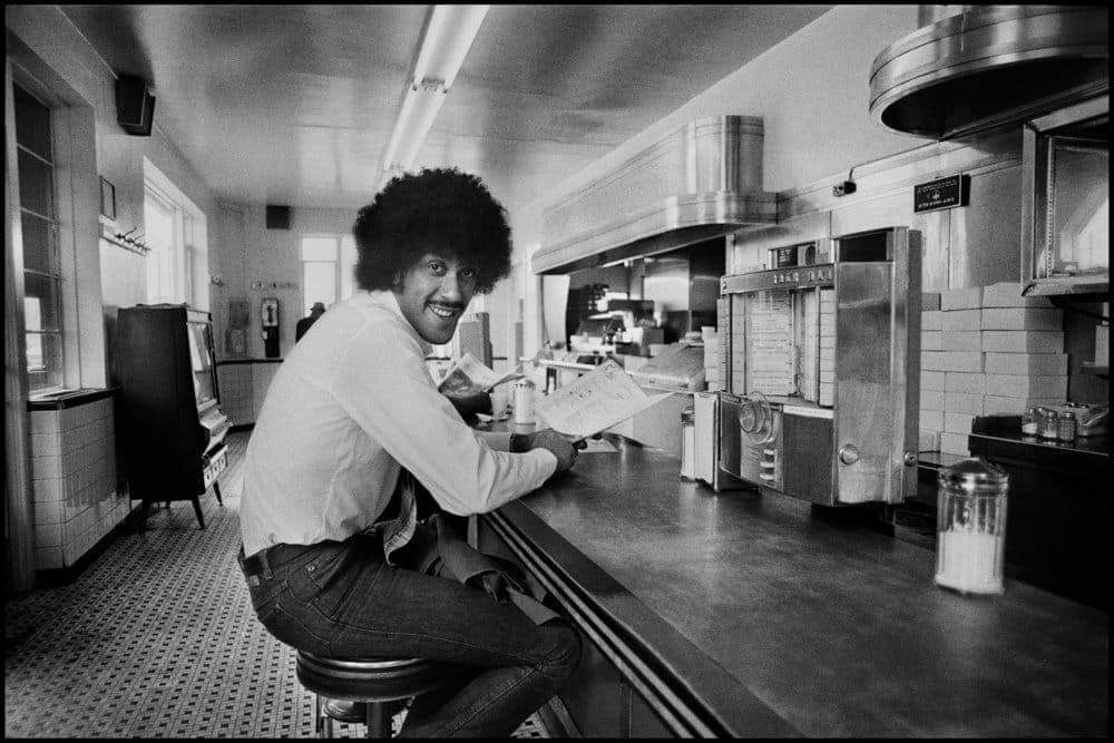 A person with an afro hairstyle sits at a retro diner counter, turning and smiling towards the camera. They hold an open magazine, with stainless steel kitchen equipment visible in the background. The ambiance is classic and nostalgic.