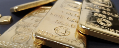 Close-up of several shiny gold bars on a dark surface. The bars are engraved with fine details, including numbers, a stamp, and the word "GOLD." The focus is on the front bar, while others are slightly blurred in the background.