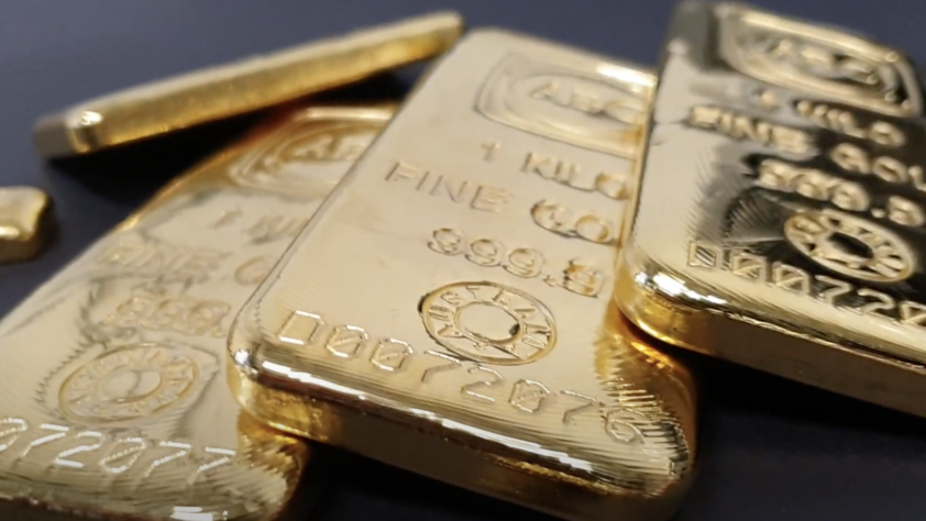 Close-up of several shiny gold bars on a dark surface. The bars are engraved with fine details, including numbers, a stamp, and the word "GOLD." The focus is on the front bar, while others are slightly blurred in the background.