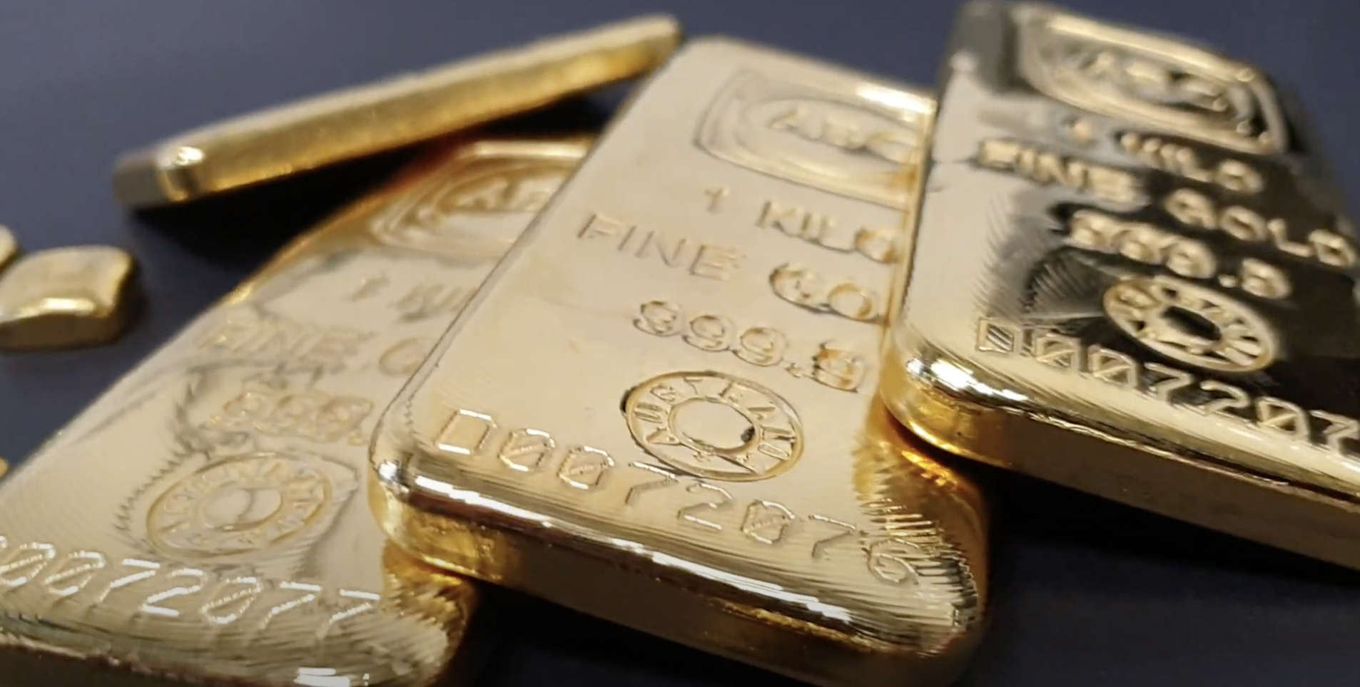 Close-up of several shiny gold bars on a dark surface. The bars are engraved with fine details, including numbers, a stamp, and the word "GOLD." The focus is on the front bar, while others are slightly blurred in the background.