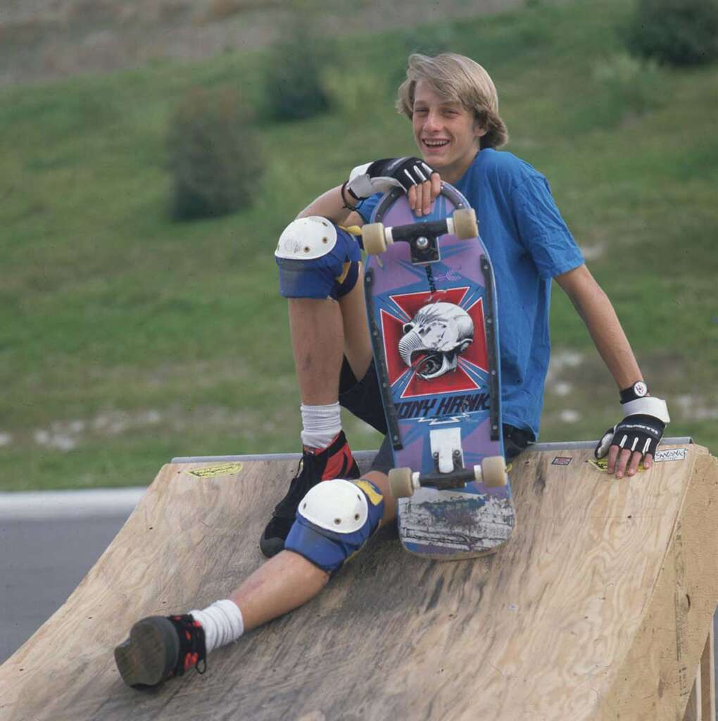 Young person sitting on a skateboard ramp, holding a skateboard. They wear a blue shirt, knee and elbow pads, and gloves. The skateboard has a graphic design on it. The background shows greenery and a clear sky.