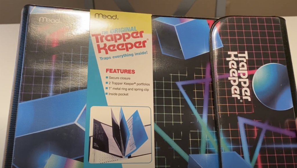 A Mead Trapper Keeper binder with a retro geometric design featuring spheres and cubes. A label highlights features: secure closure, two portfolios, a metal ring, a spring clip, and an inside pocket.