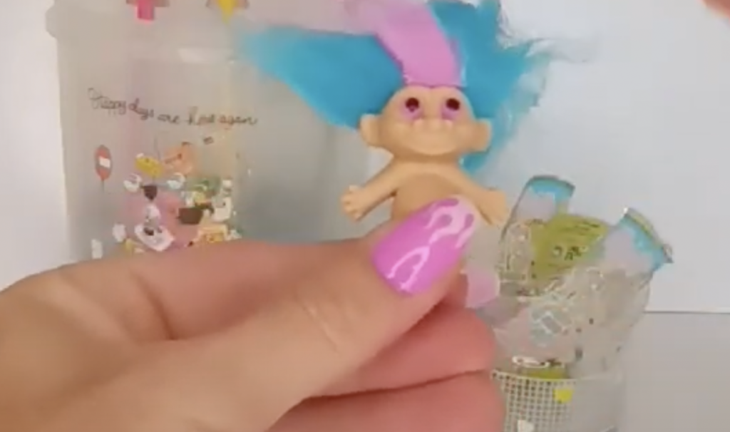 A person with vibrant purple manicured nails holds a small troll doll with bright blue hair and a pink stripe. In the background, there are decorative containers with colorful designs and text.