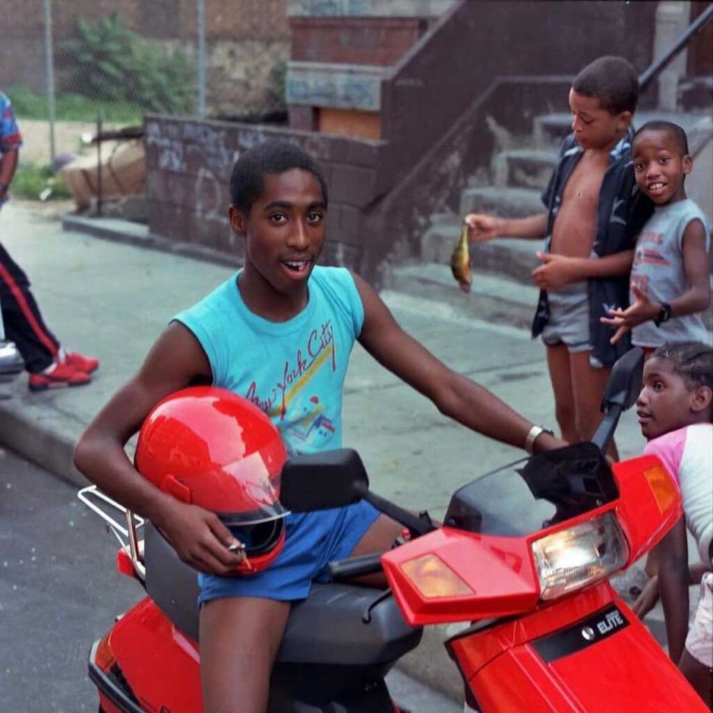 A young person smiles while sitting on a red scooter, holding a matching helmet. Three other children are in the background: one looks at the camera, one stands shirtless holding something, and the third sits on stairs. It's a sunny day in an urban setting.