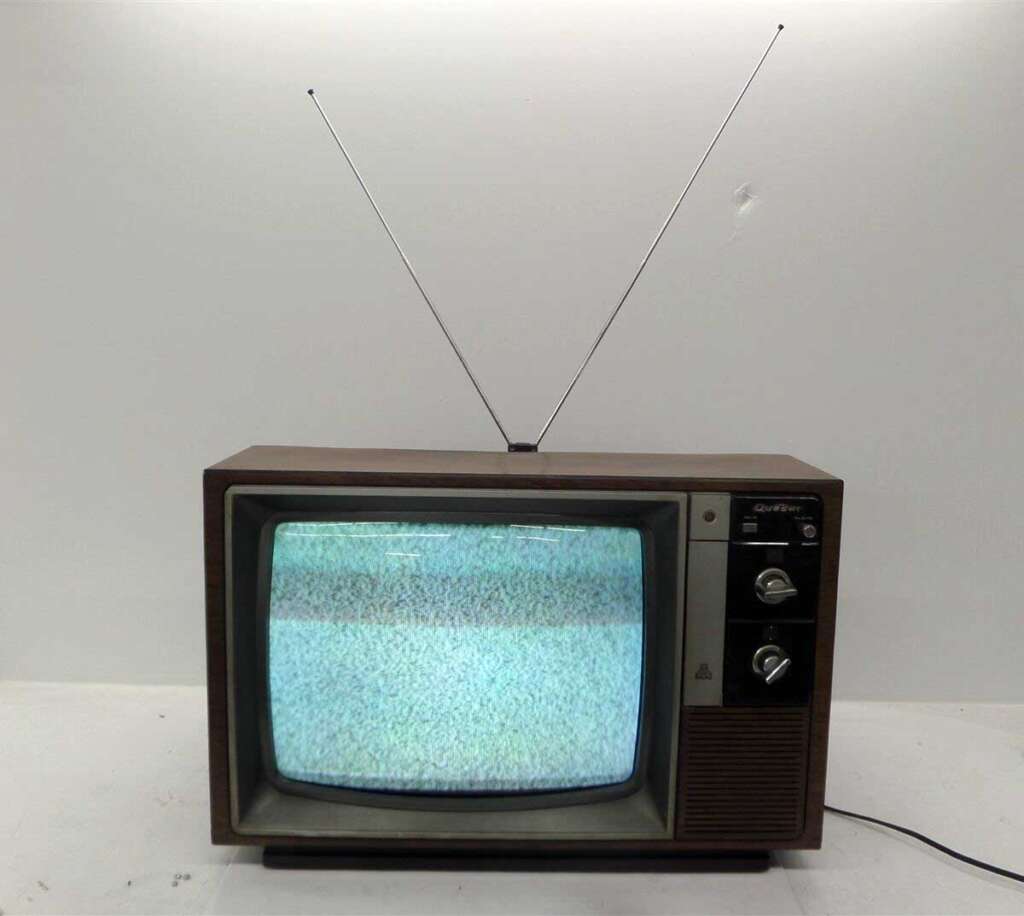 A vintage television with a wood-paneled exterior and two antenna rods displays static on its screen. It has knobs for channel and volume control on the right side. The background is a plain, light-colored wall.