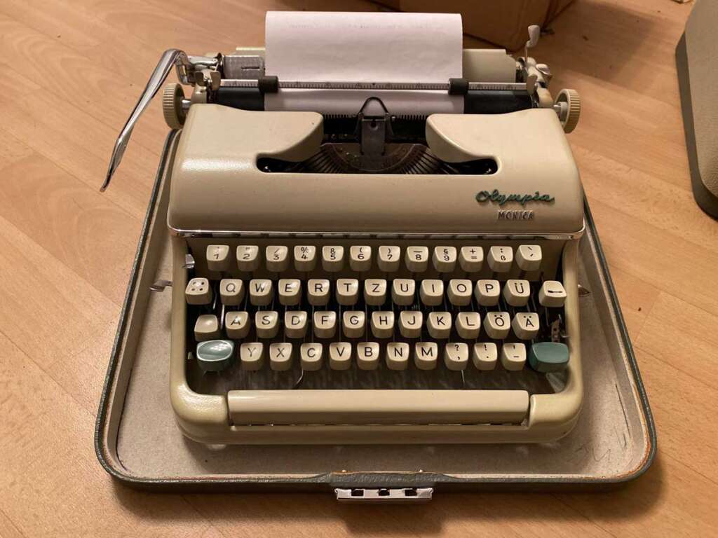 A vintage Olympia Monica typewriter with a sheet of paper inserted, placed inside an open case on a wooden floor. The keyboard features a QWERTZ layout with German characters, and the typewriter has a beige color.