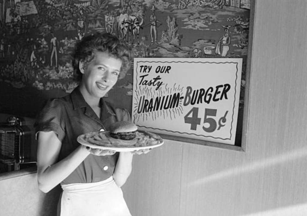 A woman smiles while holding a plate with a burger and fries. A sign in the background reads, "Try our tasty uranium burger 45¢." The setting appears to be a vintage diner with a decorative mural.