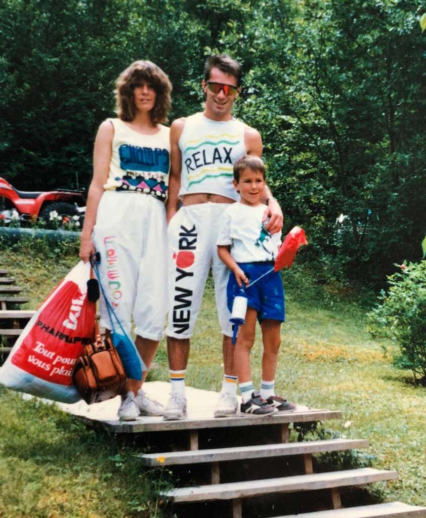 A family of three stands on wooden steps outdoors. The woman wears a sleeveless top and white pants. The man sports a sleeveless "RELAX" shirt and New York pants. The child holds a red bucket. Trees and greenery are in the background.