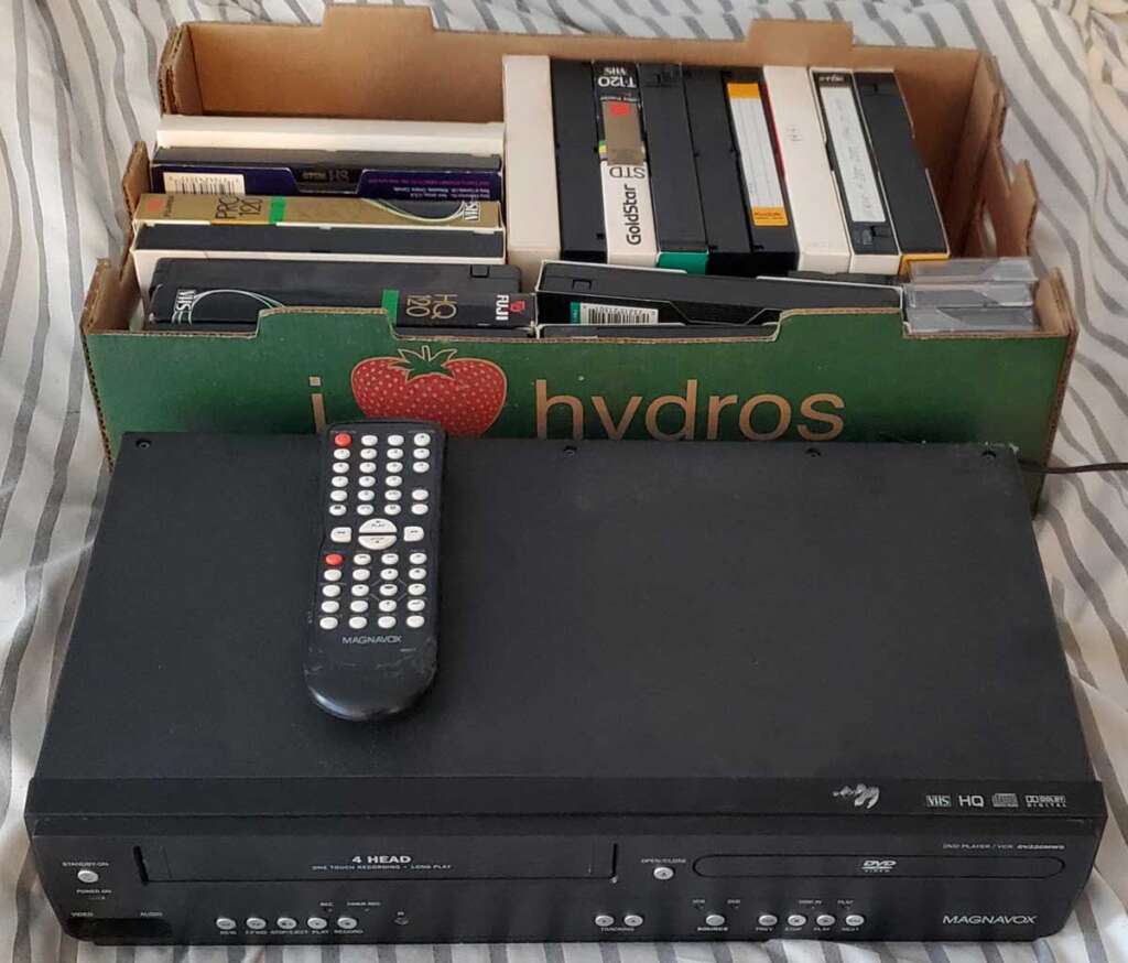 A VHS player with a remote is in front of a cardboard box filled with various VHS tapes. The box has "hydros" written on it with an illustrated apple. It's resting on a striped fabric surface.