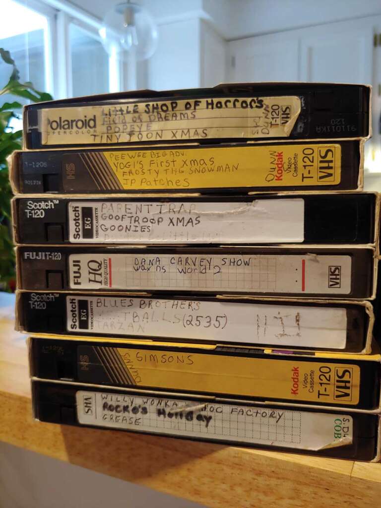 A stack of eight VHS tapes labeled with various movie and TV titles, including "Little Shop of Horrors," "Carpenters," "The Blues Brothers," "Simpsons," and "Grease." The tapes are different brands, like Polaroid and Kodak, resting on a wooden surface.